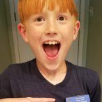 Smiling child with red hair and open mouth, wearing a dark shirt indoors