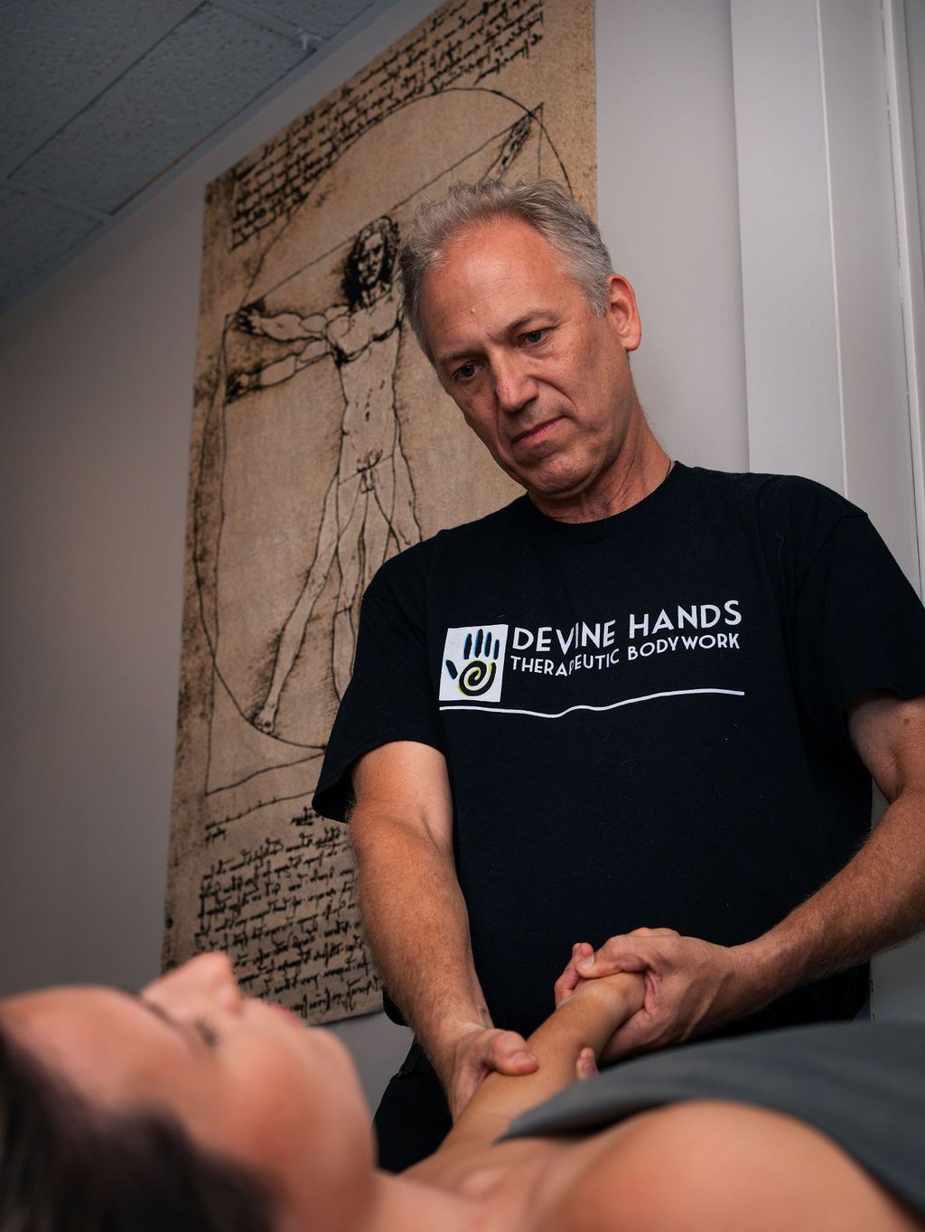 Man standing over a person lying down, holding their hand in a clinic room with a wall diagram.