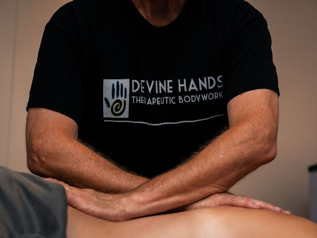 Person in a black “Divine Hands Therapeutic Bodywork” shirt resting crossed arms on a massage table