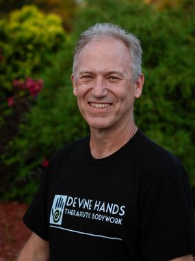 Smiling man in black DEAF HANDS T-shirt standing outdoors with greenery and flowers behind him