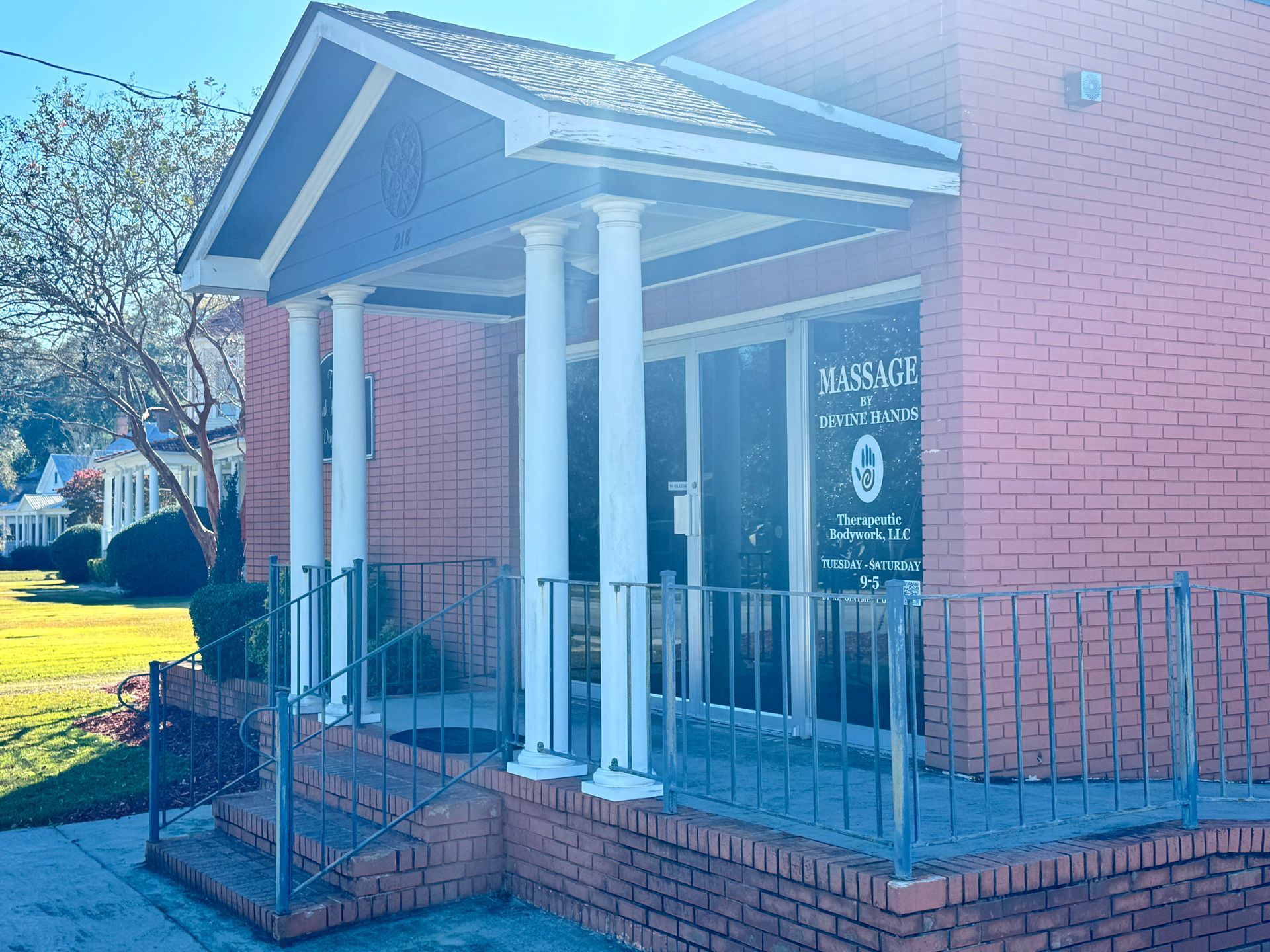 Brick building entrance with white columns, metal railing, and a glass door in bright sunlight