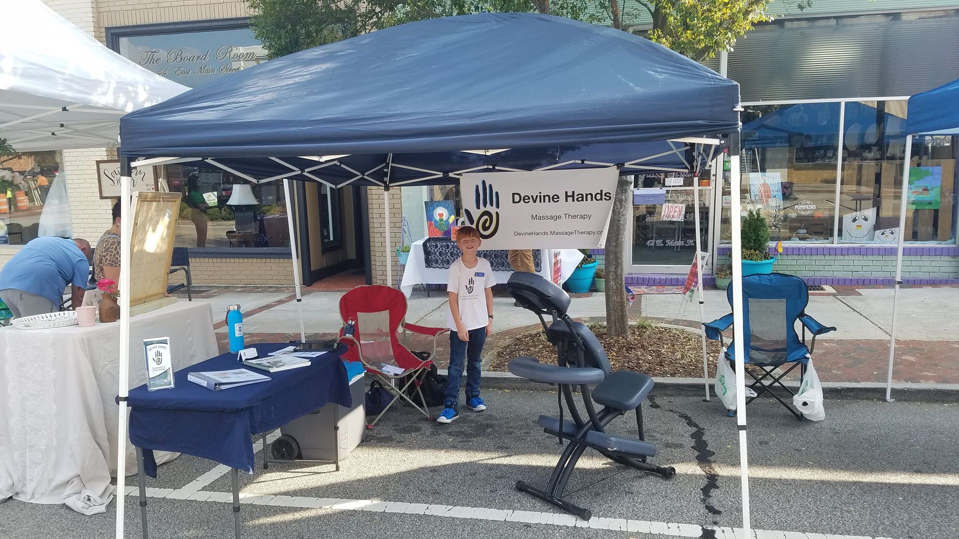 Blue canopy tent with folding tables, chairs, and people at an outdoor street fair booth
