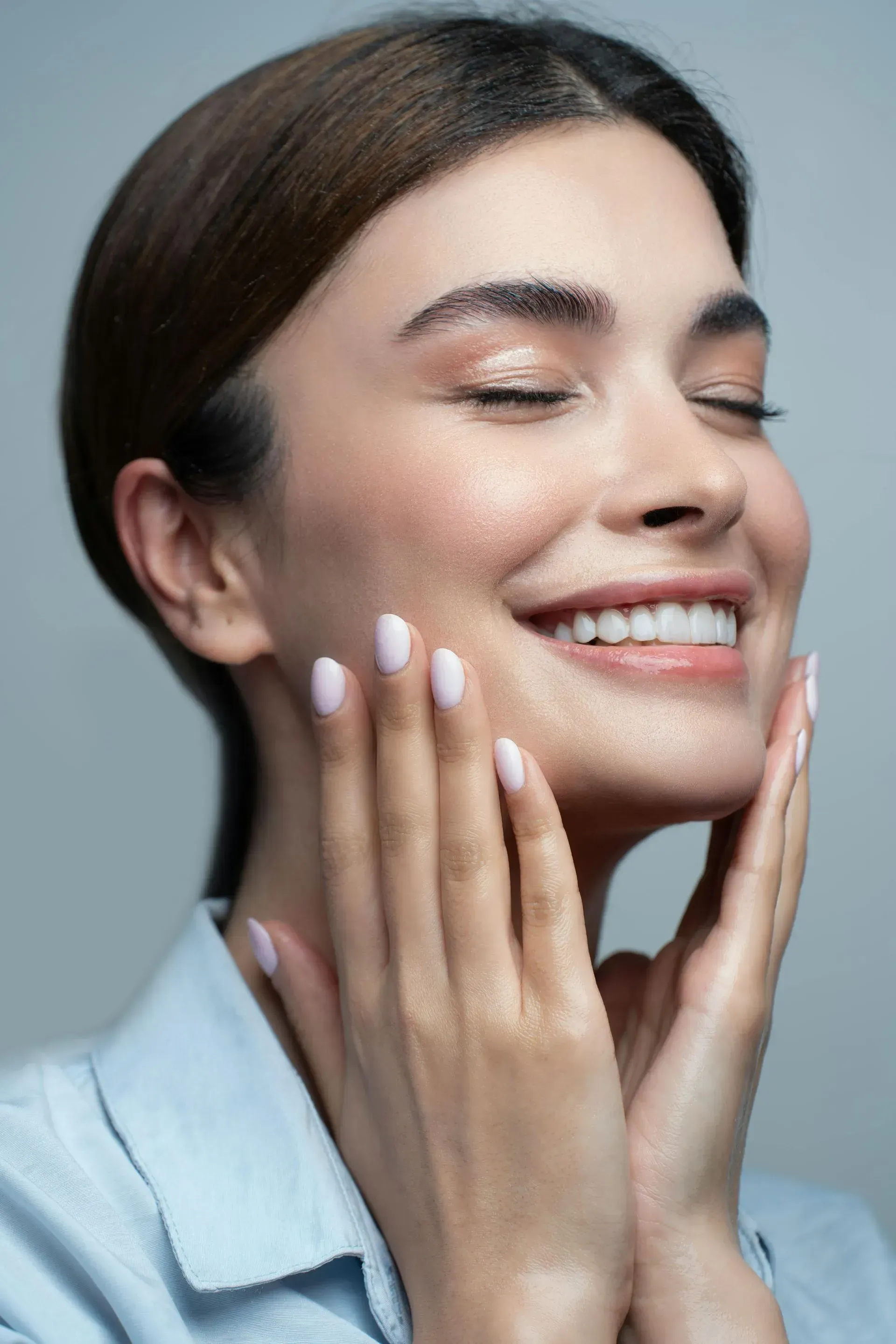 Smiling person with eyes closed gently touching their cheeks against a gray background