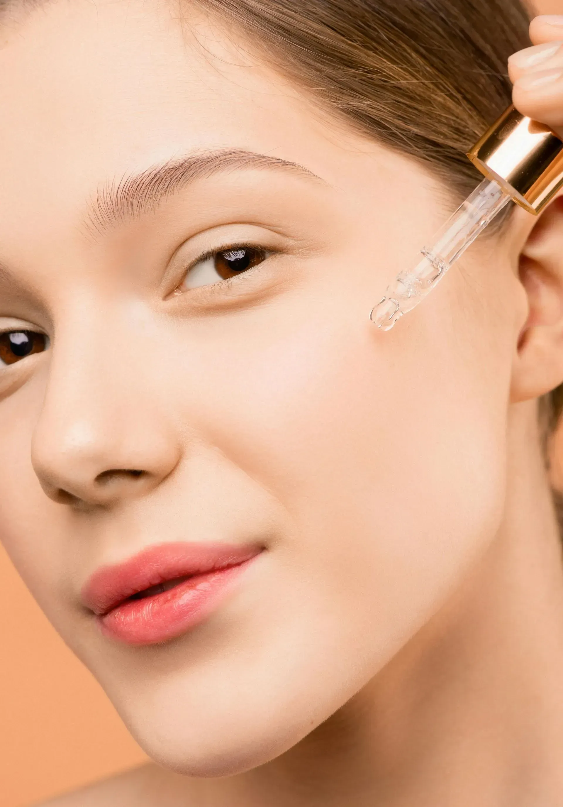 Woman applying serum to her cheek with a dropper, close-up against a peach background