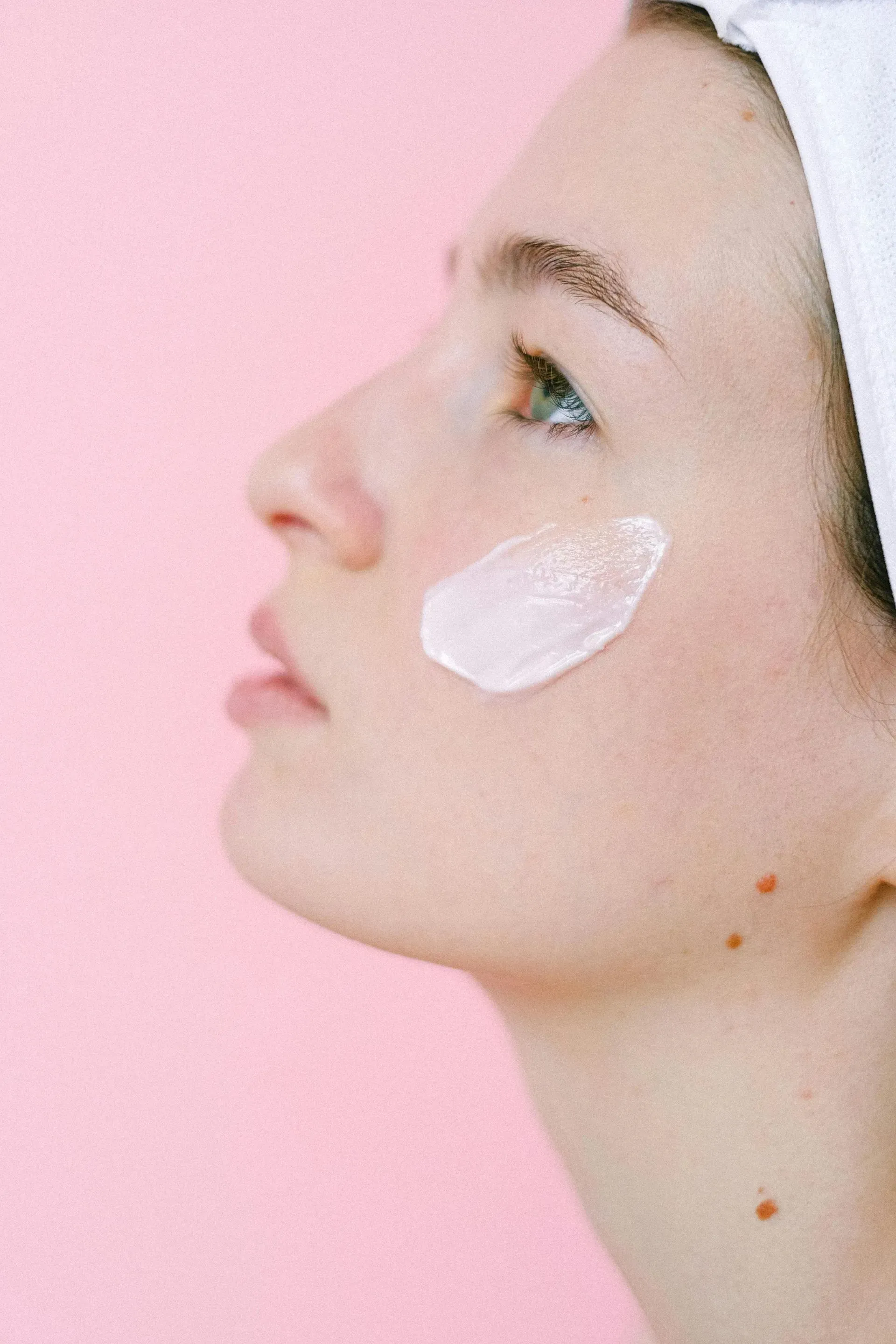 Profile of a woman with a white facial cream patch on her cheek against a pink background
