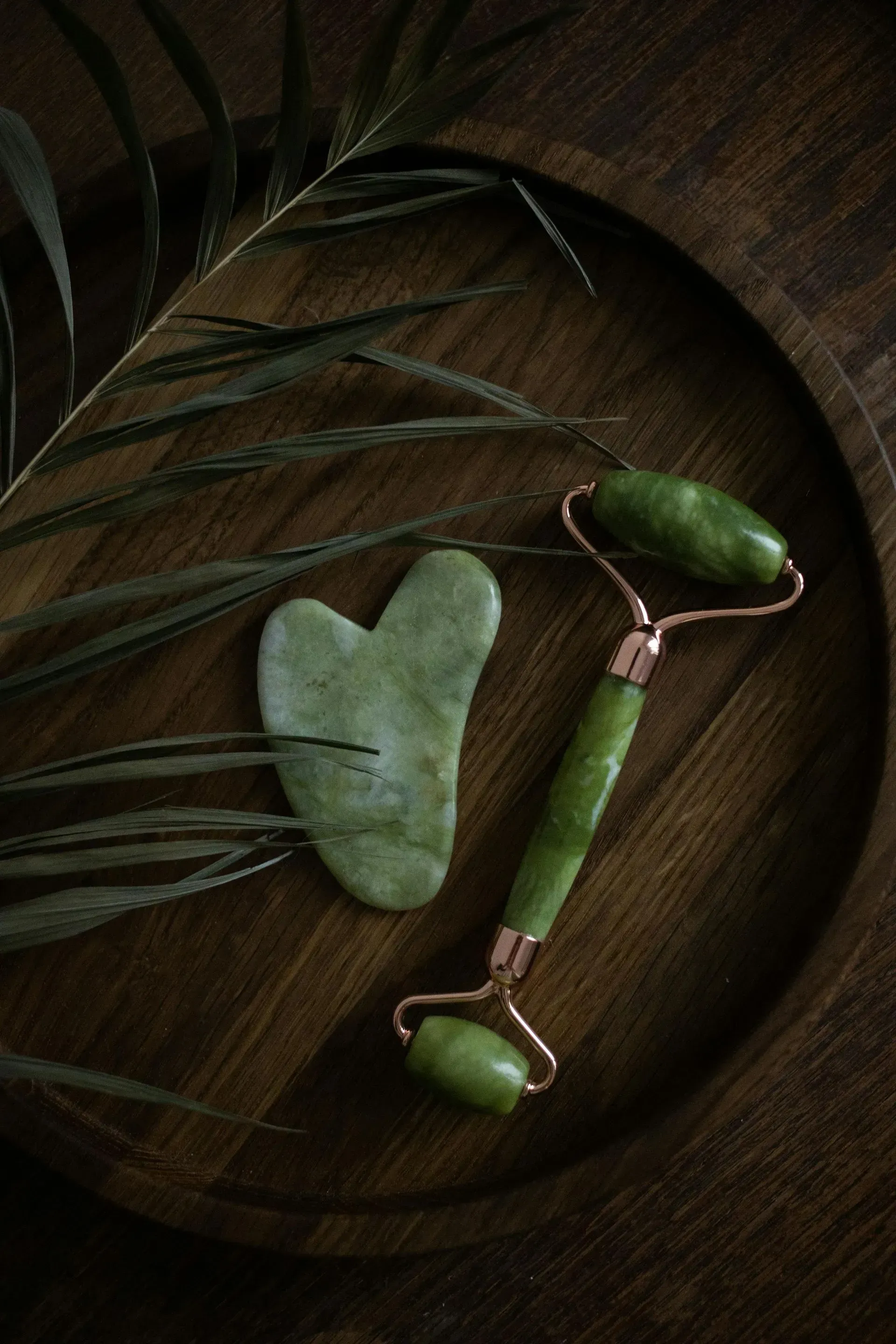 Green jade facial roller and heart-shaped gua sha on a dark wooden tray with leaves