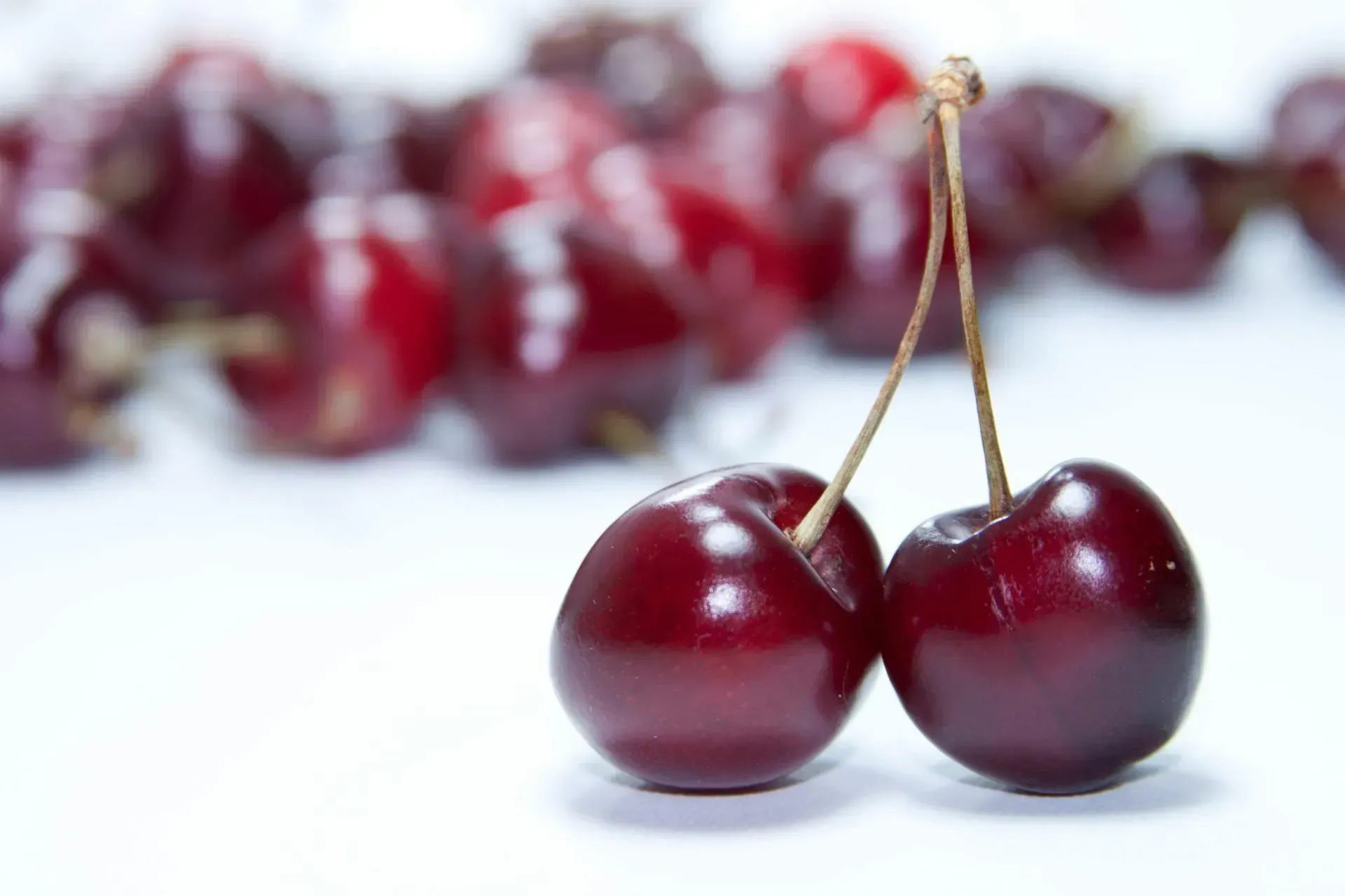 Two dark red cherries with stems on a white surface, with more cherries blurred in the background