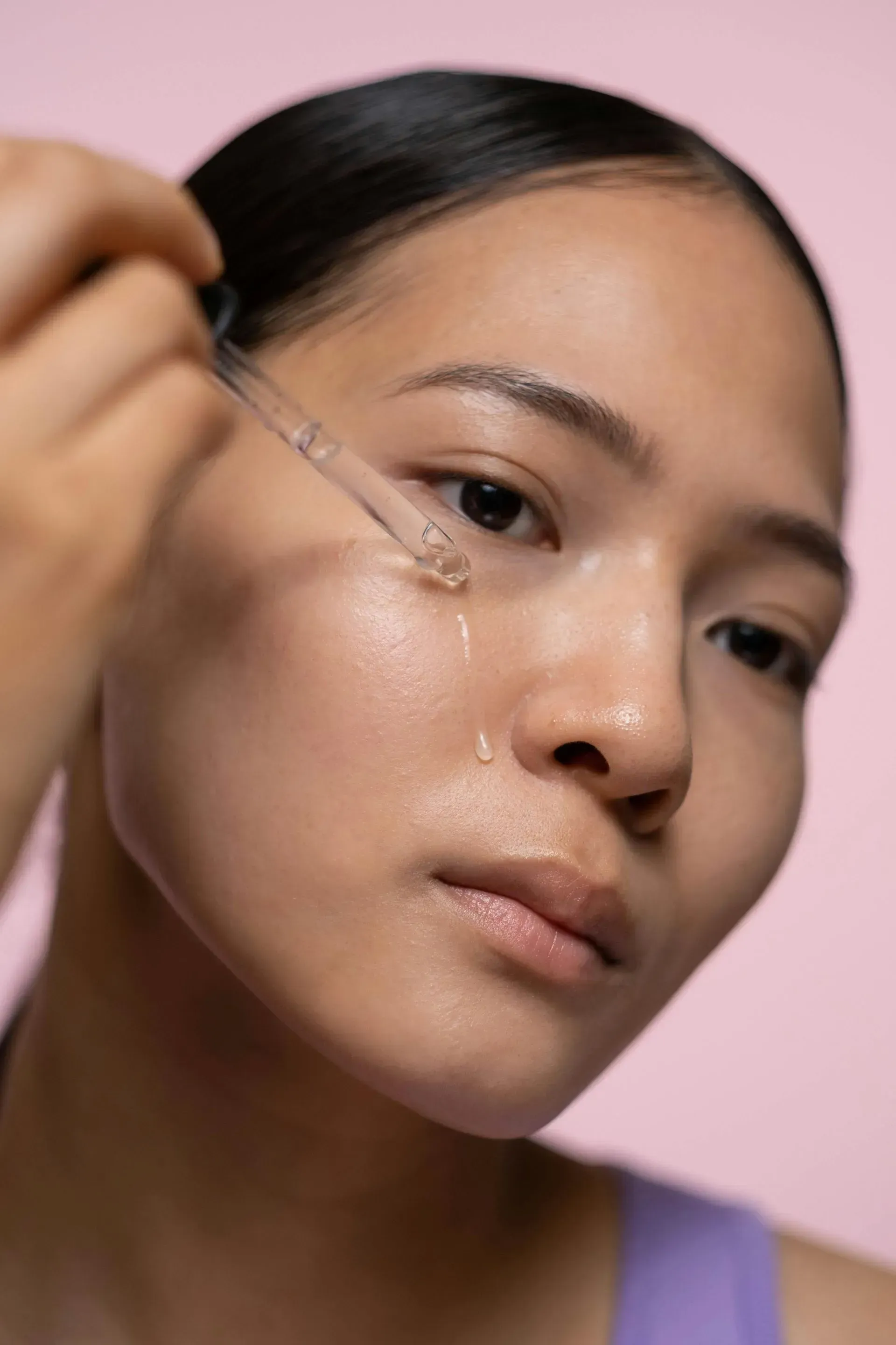 Woman applying facial serum with a dropper against a pink background.