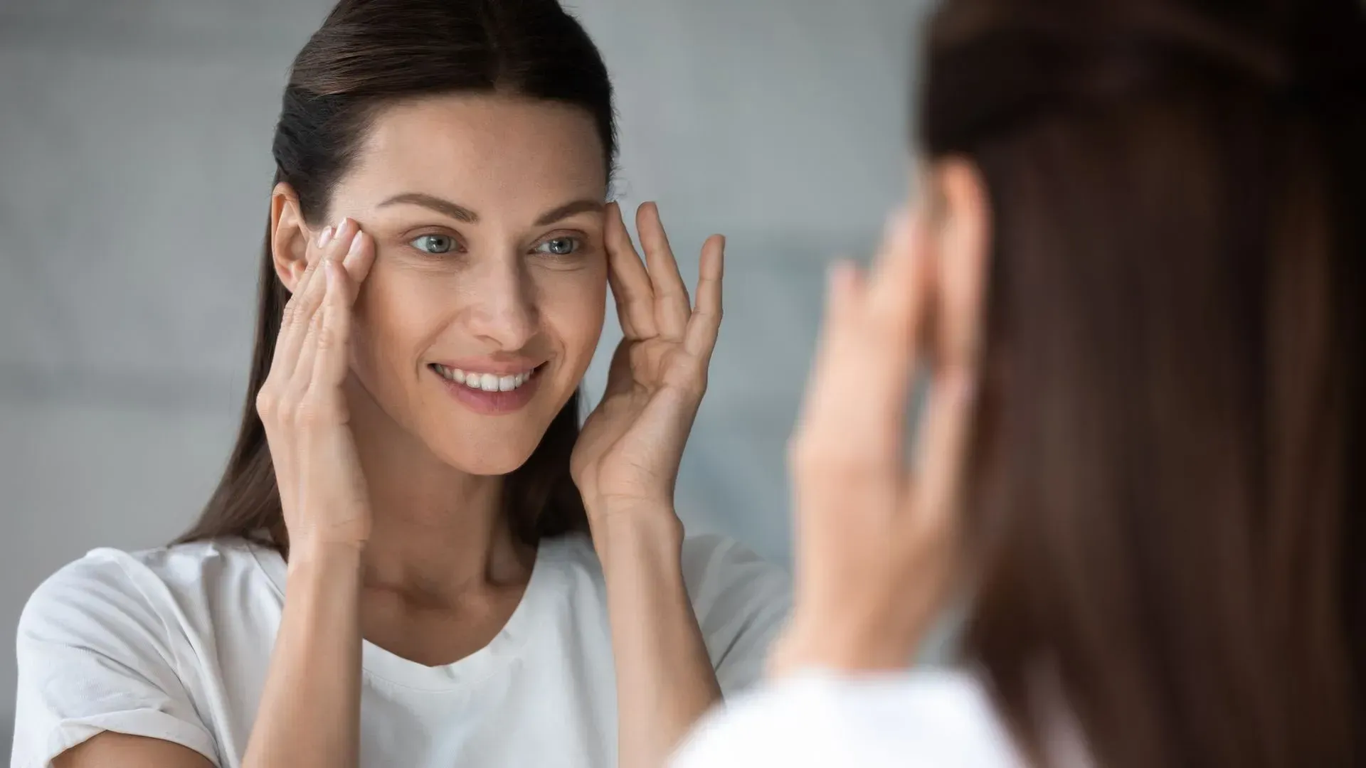 Woman smiling at a mirror and touching her face in a bright room