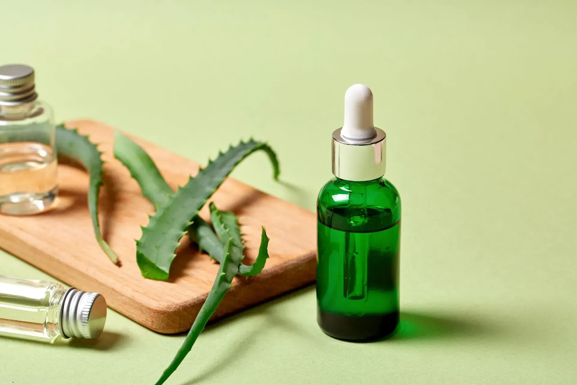 Green essential oil bottle with aloe vera leaves on a wooden board, with small glass vials nearby
