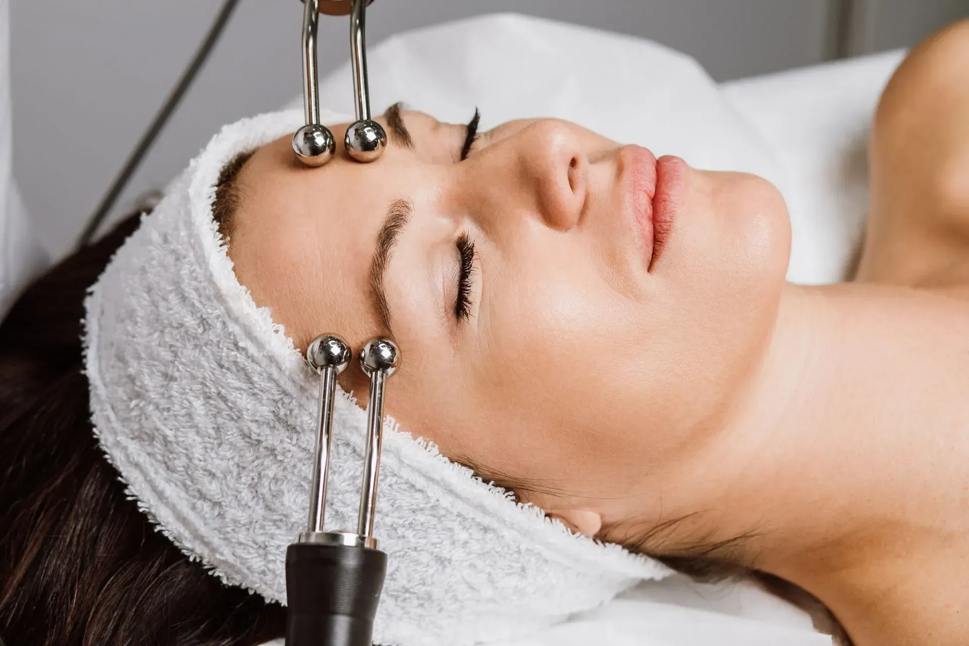 Woman receiving a facial treatment with probes on her forehead, lying on a spa bed with a towel wrap
