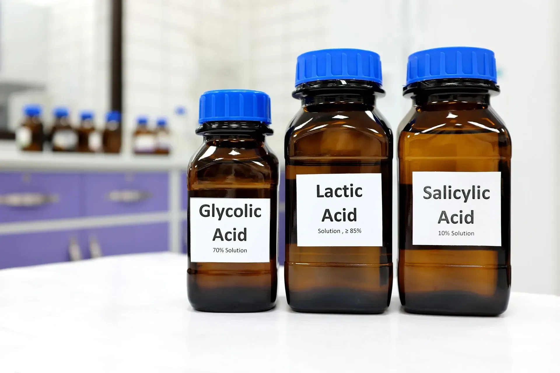 Three brown bottles labeled glycolic acid, lactic acid, and salicylic acid on a lab counter