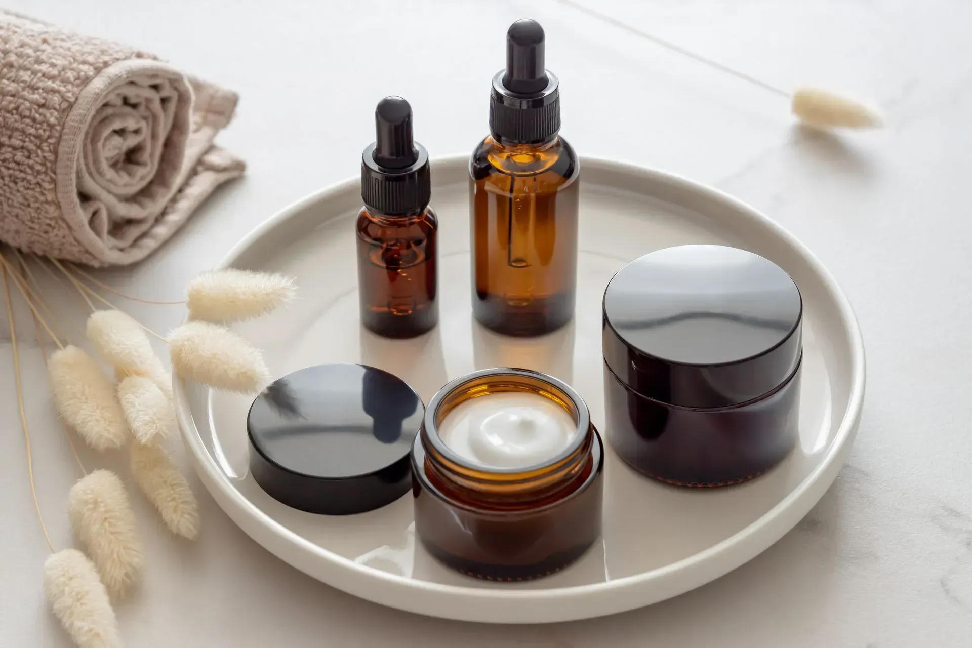 Amber skincare serum bottles and cream jars on a white tray with dried flowers