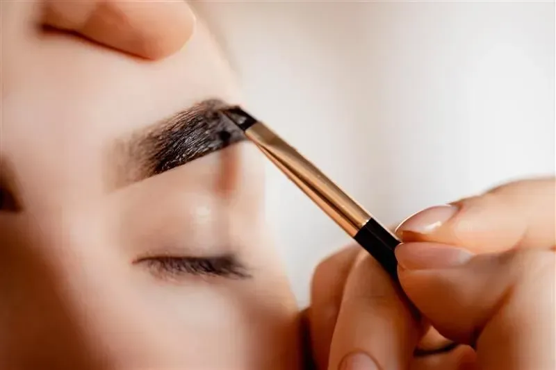 Hand applying dark eyebrow makeup with an angled brush to a closed eye