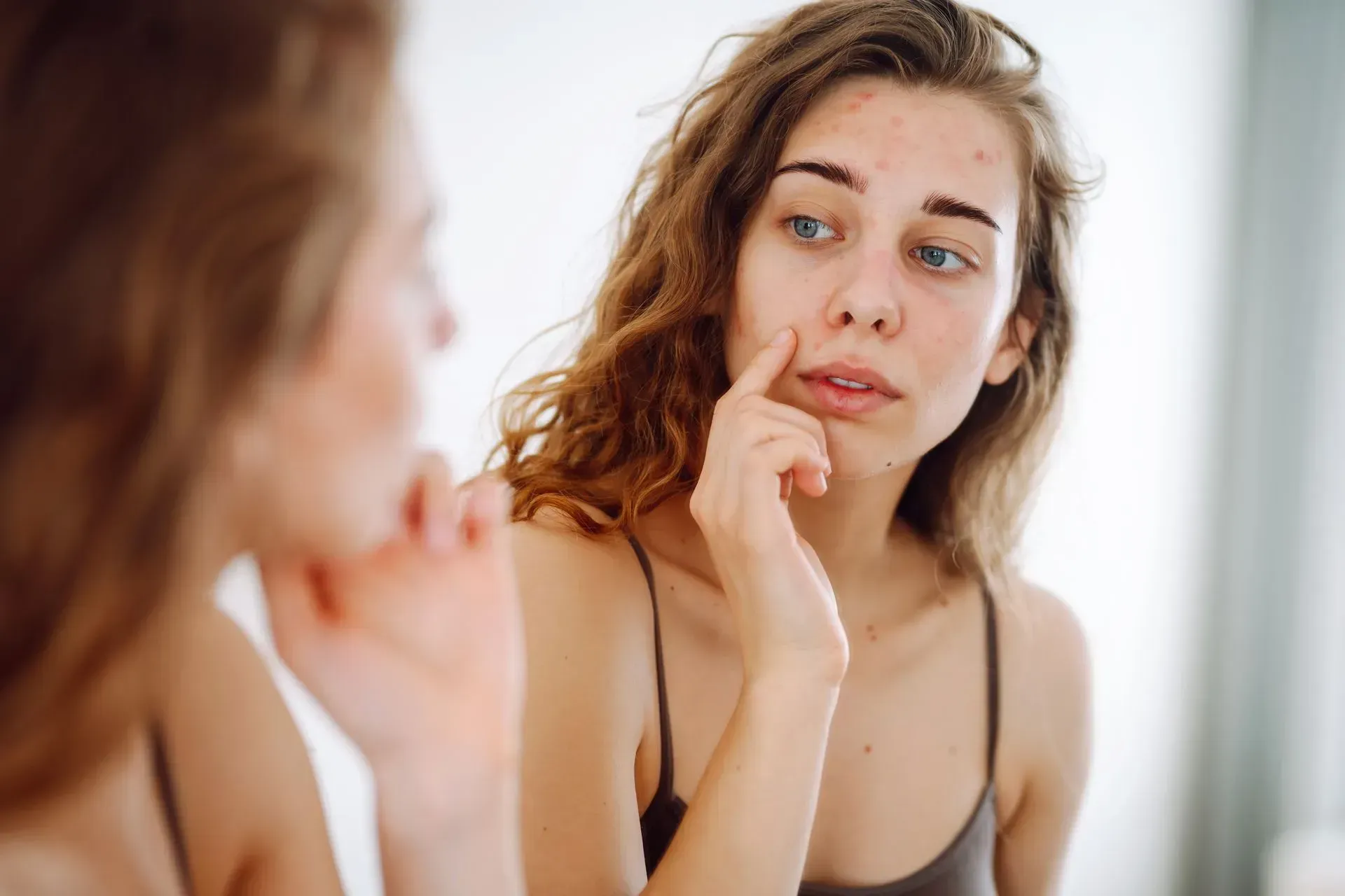 Woman examining her face in a mirror, touching a blemish on her cheek.