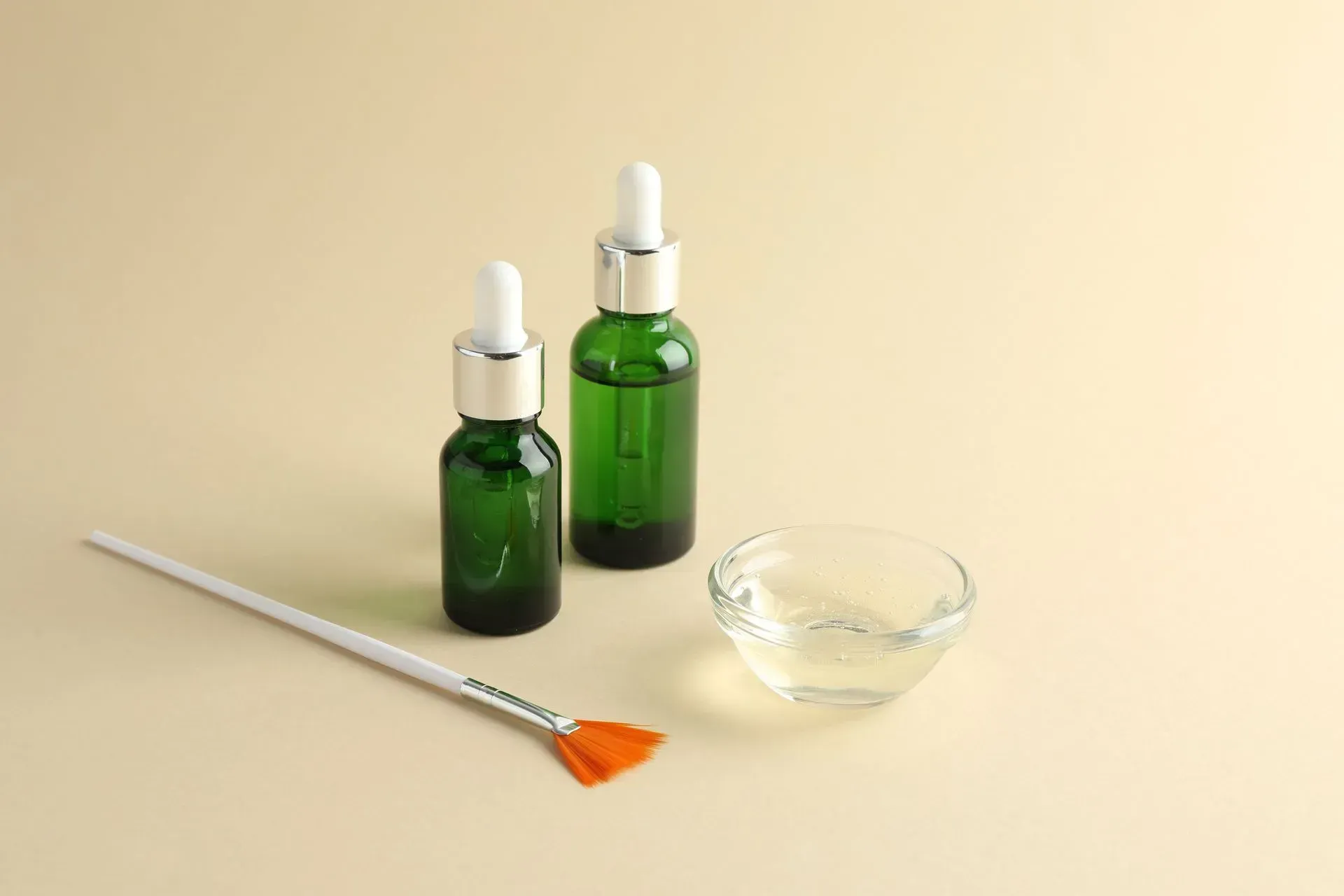 Two green dropper bottles, a glass bowl, and a paintbrush on a beige surface