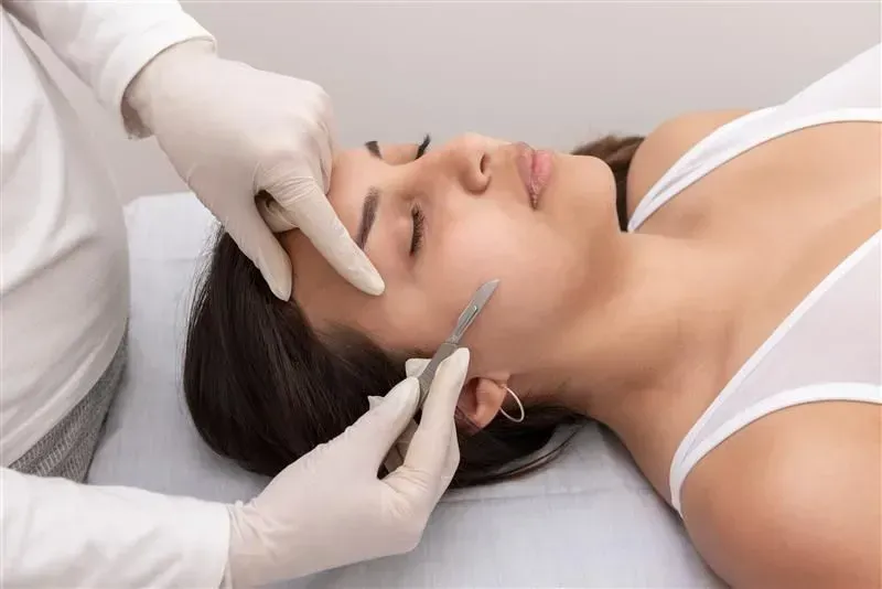 Gloved practitioner using a syringe near a woman’s eyebrow during a cosmetic procedure