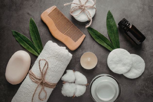Spa essentials on gray fabric: soap, loofah, comb, cotton pads, cotton, candle, bottle, bowl, and green leaves