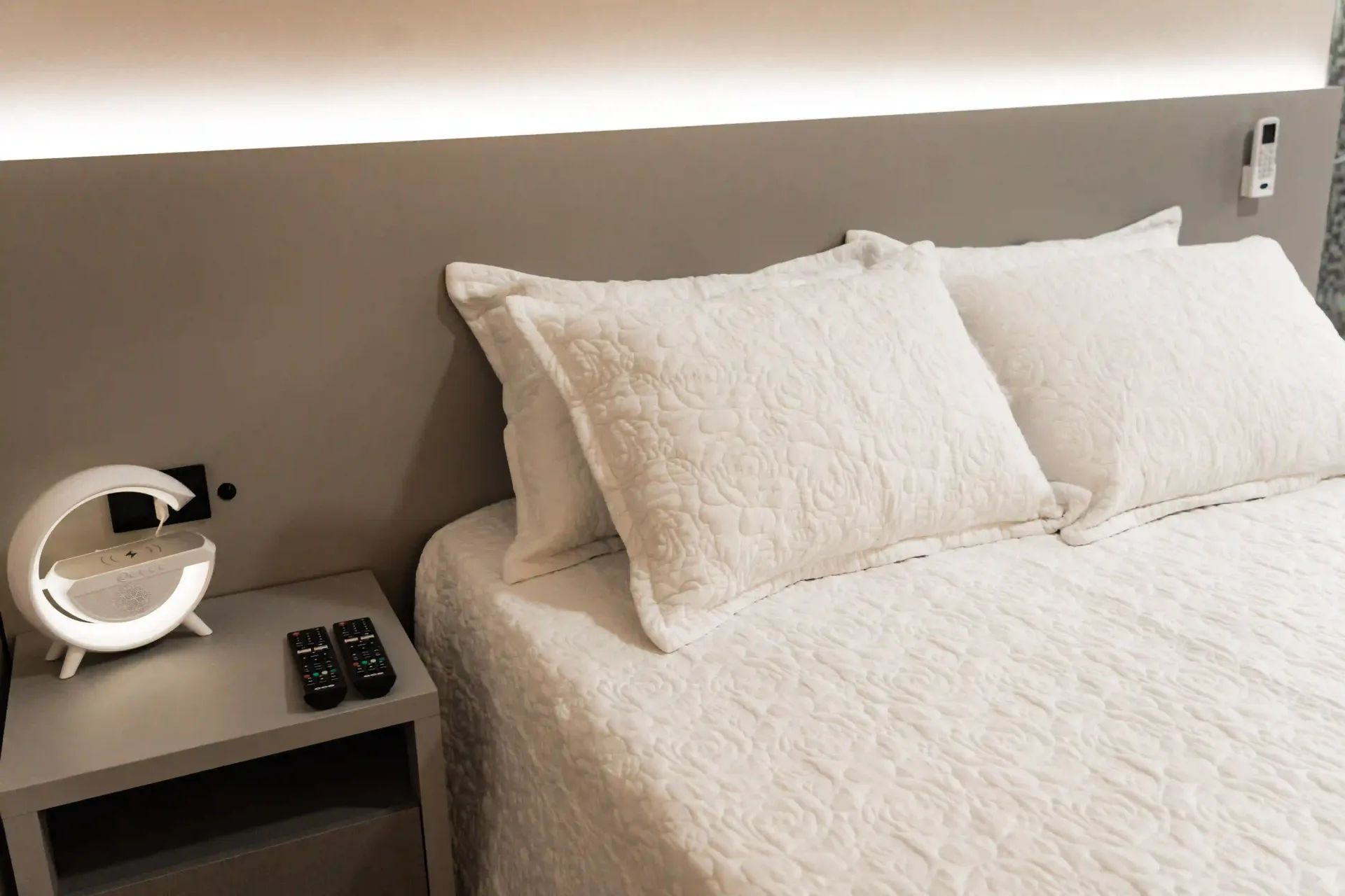 Uma cama com lençóis e travesseiros brancos e uma mesa de cabeceira com dois controles remotos MD Recanto Hotel