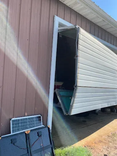 Damaged white garage door hanging open on a pink-sided building, with a solar panel leaning nearby.