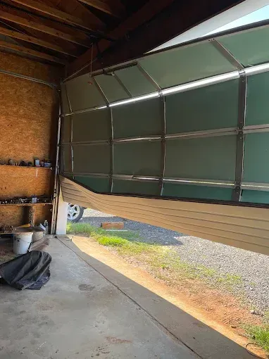 Inside a garage, a partially open green sectional door reveals sunlight and gravel outside.