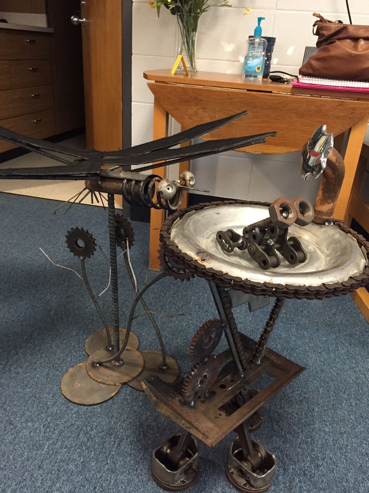 A rustic metal sculpture of a person at a sink, made from tools and scrap parts in a room.
