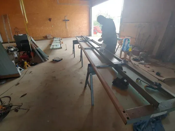 Person working in a cluttered garage workshop with long metal beams and tools on the floor.