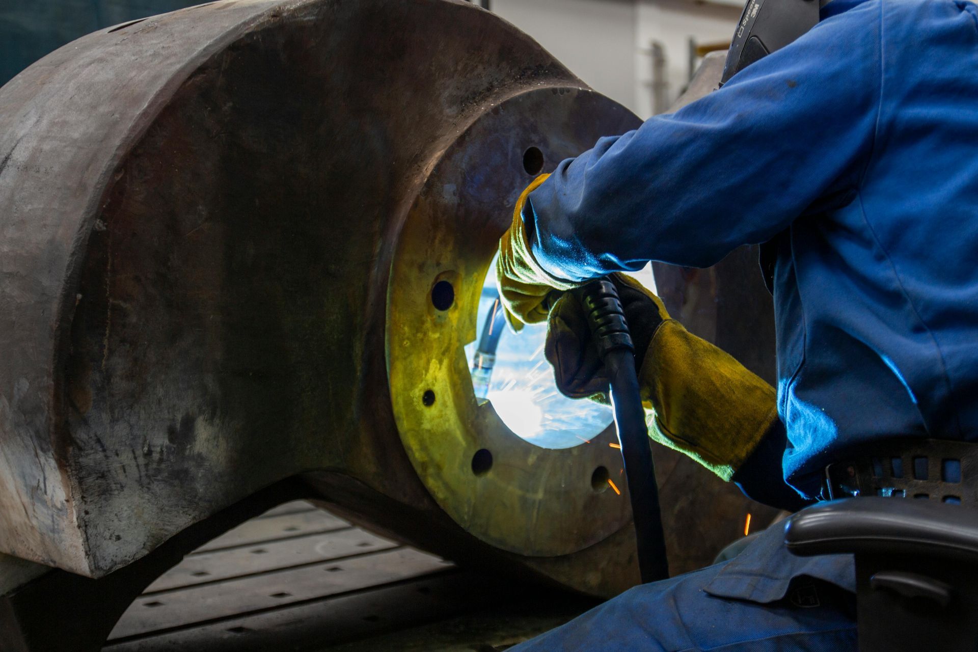 Worker in blue protective gear welding inside a large metal pipe, bright sparks illuminating the interior