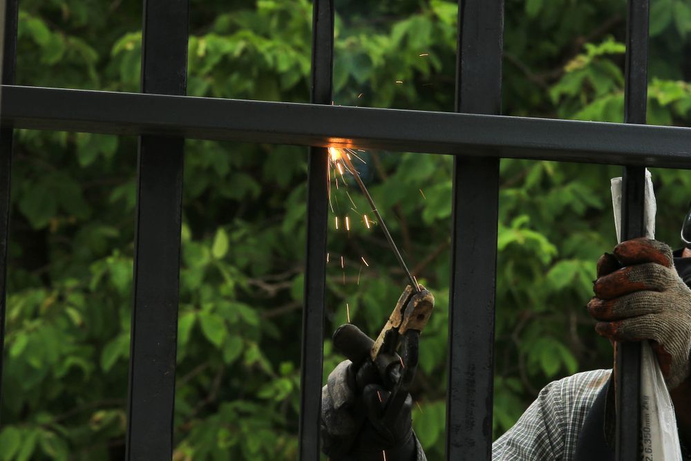 Person welding a metal fence with sparks flying near green foliage