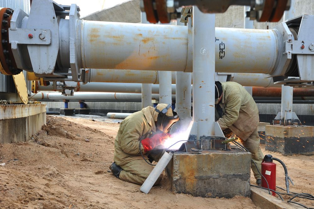Workers welding large steel pipes at a construction site, with sparks and heavy equipment in the background.