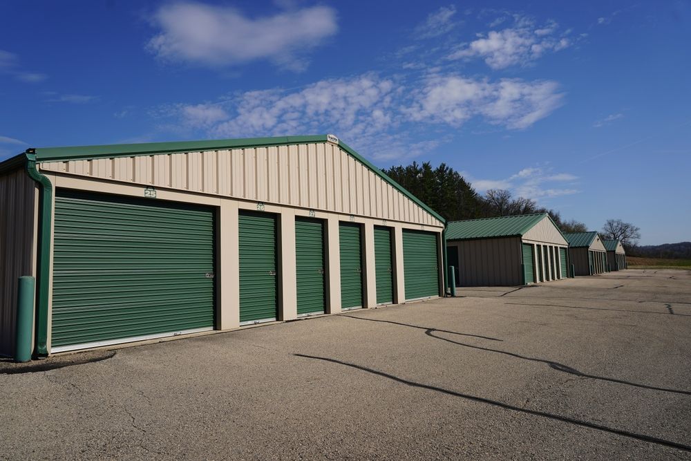 Row of green storage units with beige roofs beside a cracked paved lot under a blue sky