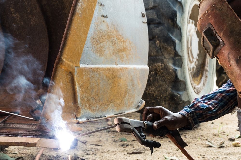 Worker welding a rusty metal vehicle frame, with bright sparks and smoke in a workshop or garage.