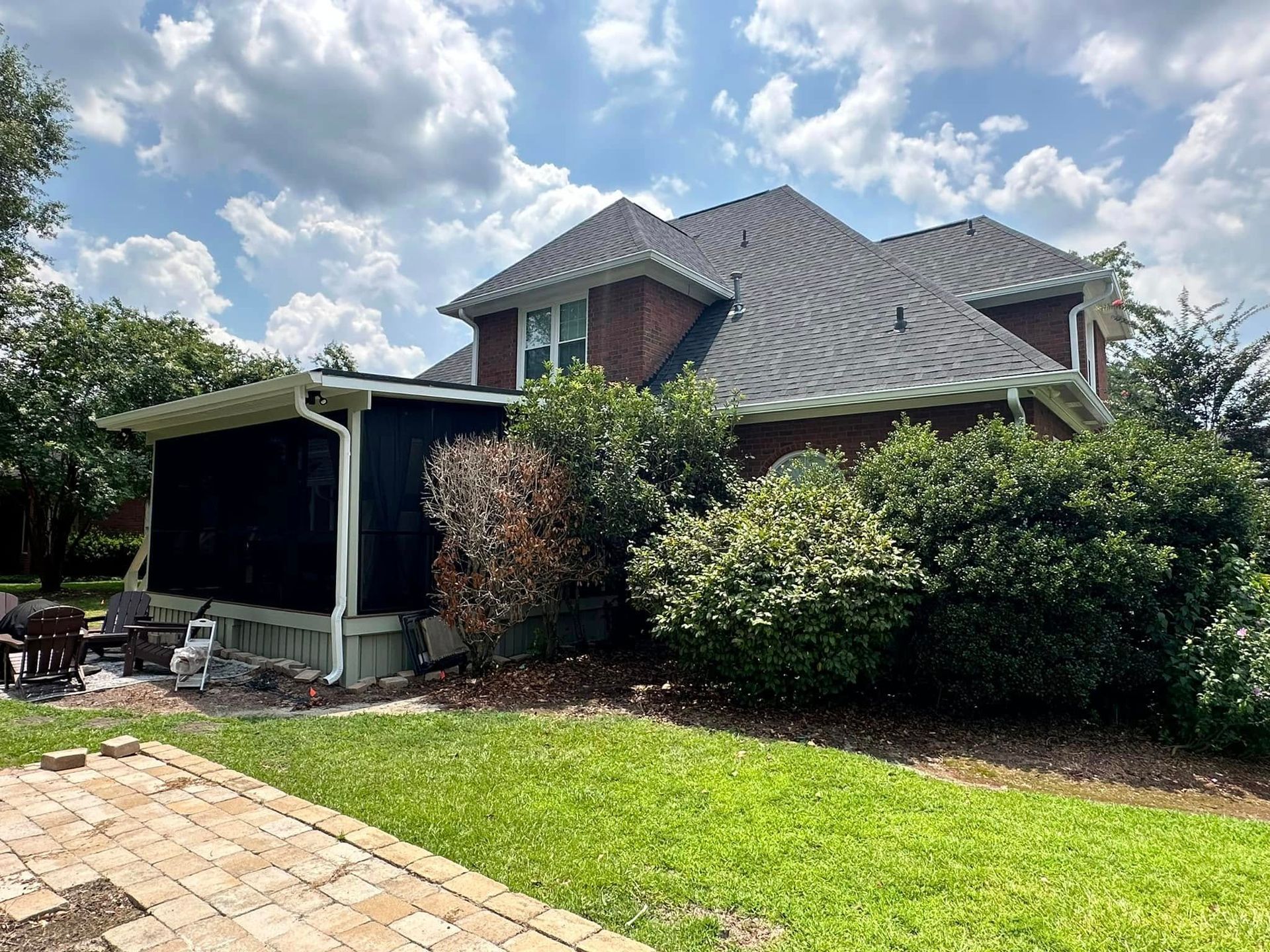 Suburban brick house with dark roof, covered patio, and green lawn under a partly cloudy sky