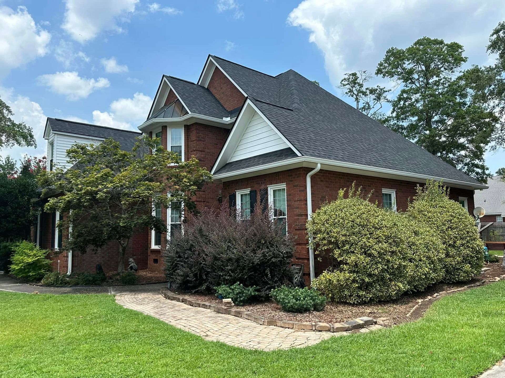 Brick suburban house with dark roof, green lawn, and trees under a partly cloudy sky
