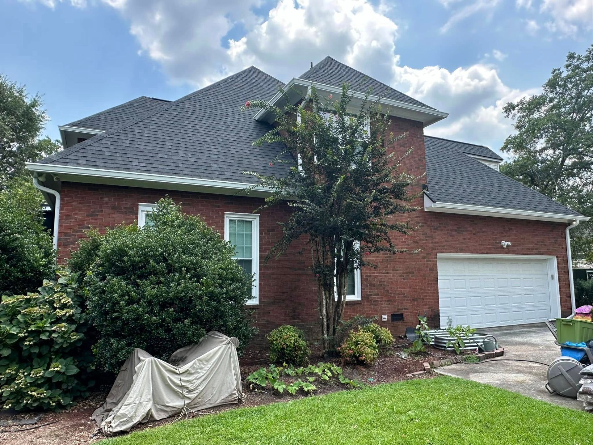 Red brick house with a gray roof, white garage, and overgrown front yard under a partly cloudy sky