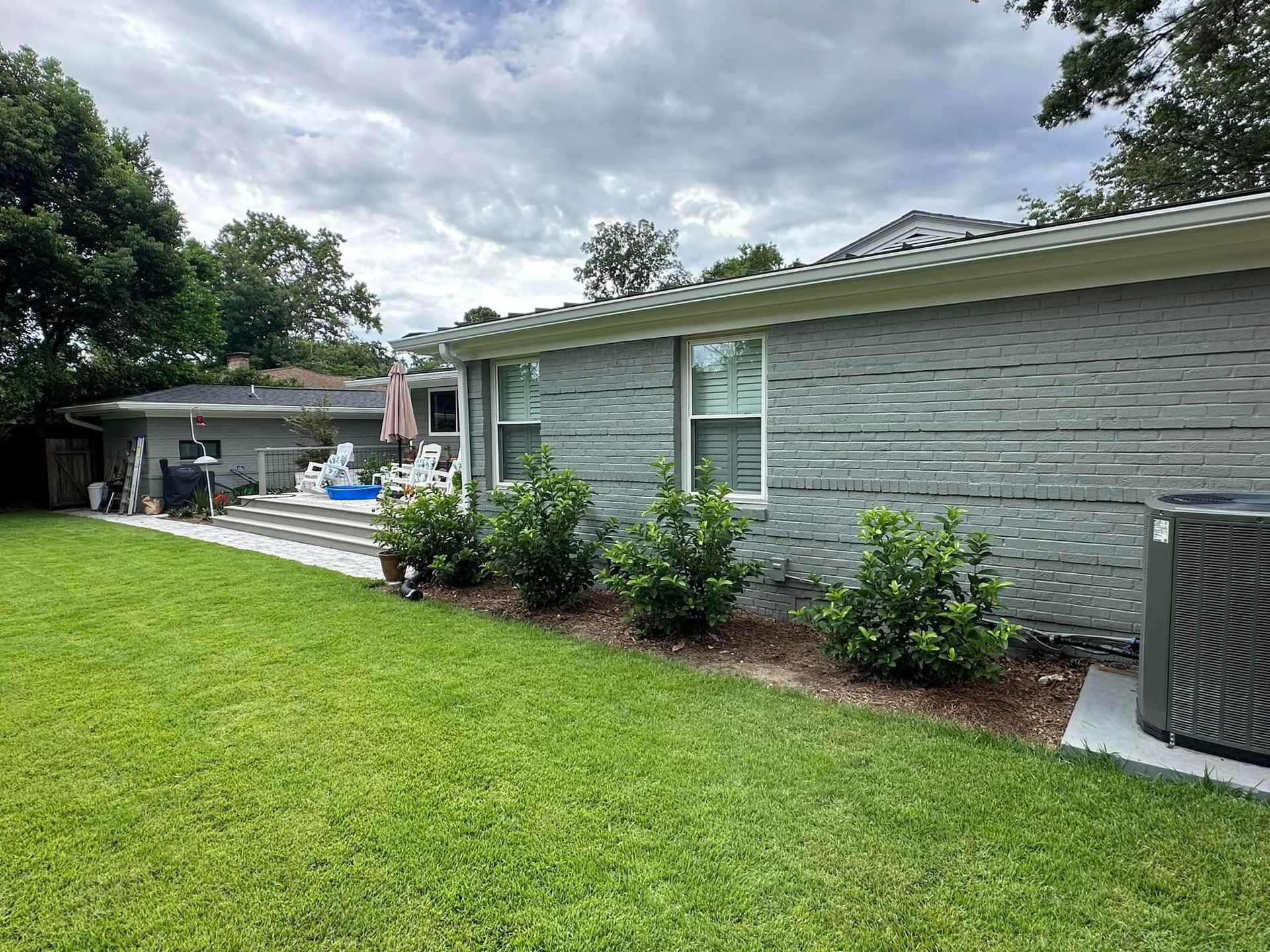 Backyard with green lawn, gray brick house, and a patio under cloudy skies
