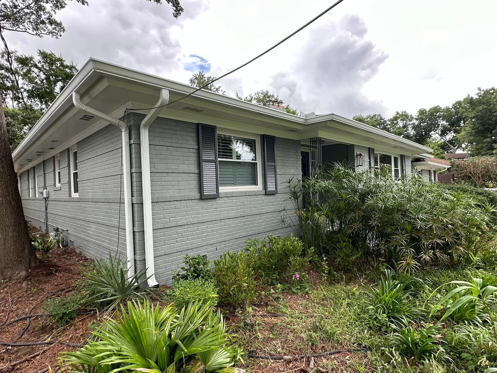 Gray brick house with white trim and shutters, surrounded by overgrown greenery under a cloudy sky