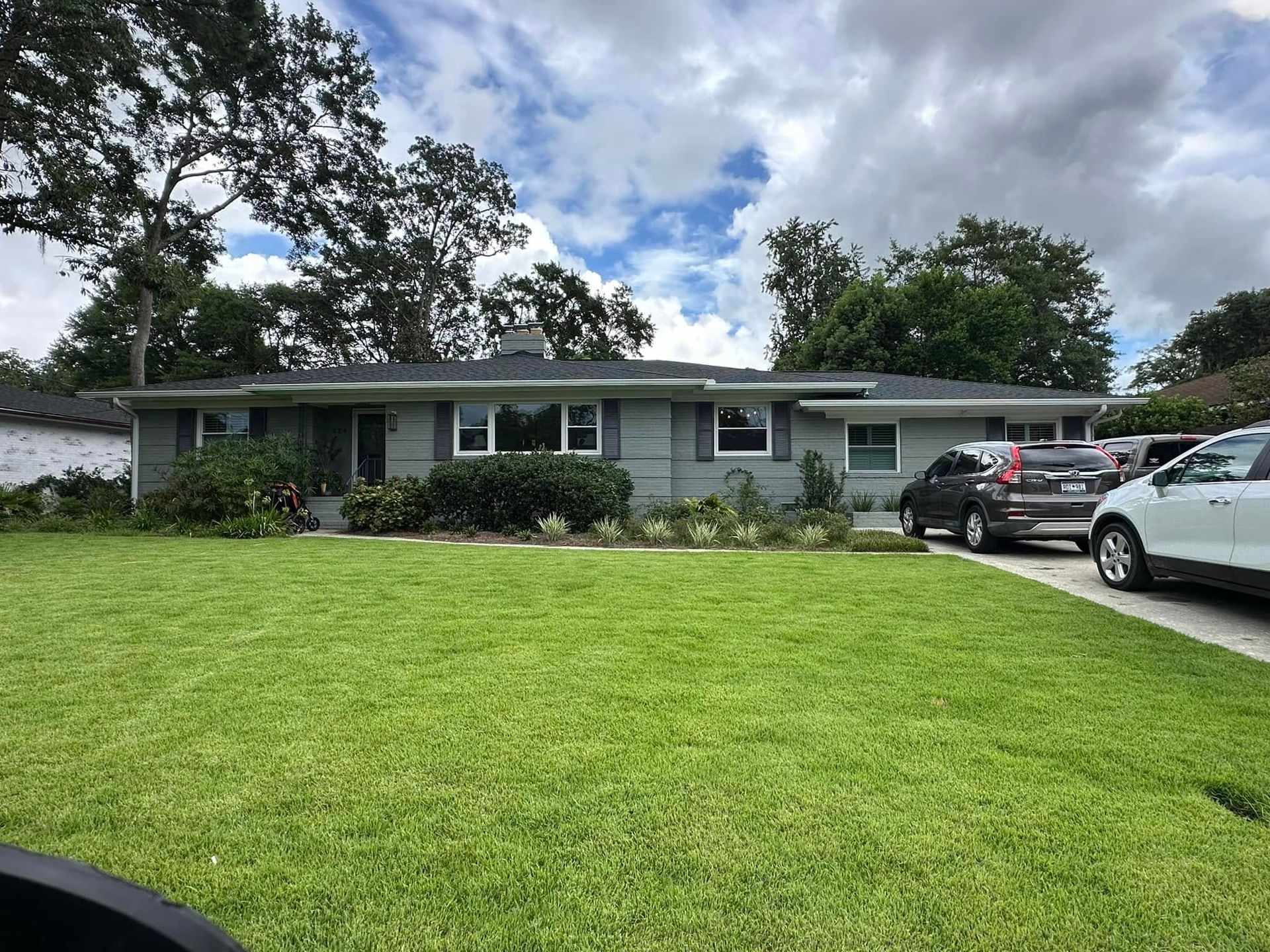 Single-story suburban house with a green lawn, cloudy sky, and two cars parked in the driveway