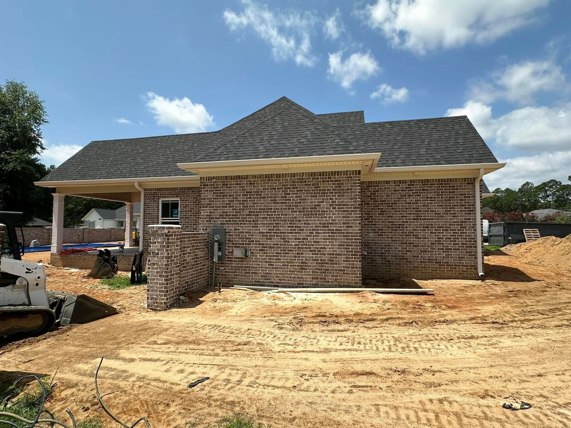 New brick house under construction with a dark shingled roof and dirt yard under a blue sky