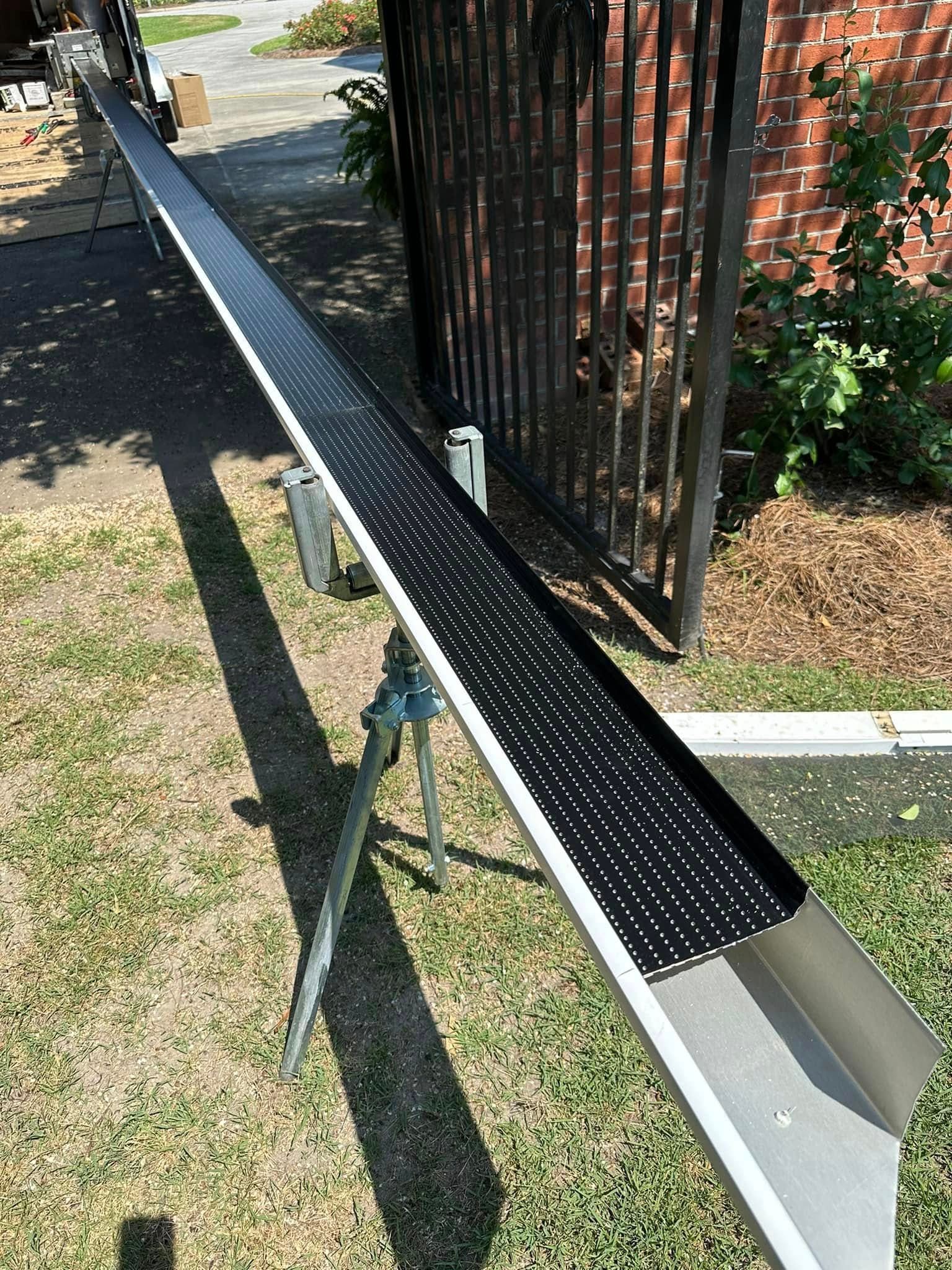 Metal ramp with black perforated surface beside a fence, leading over a small drop in a sunny yard.