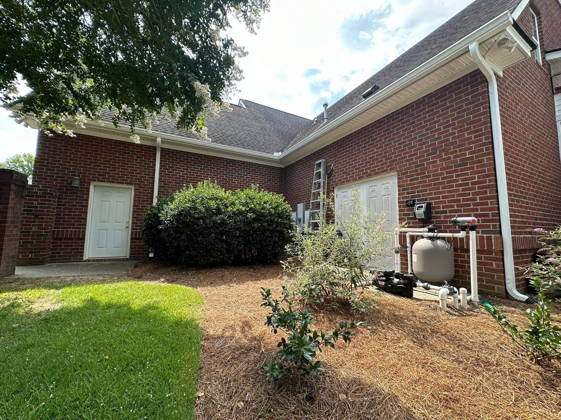 Brick house exterior with white doors, green lawn, and shrubs under a tree