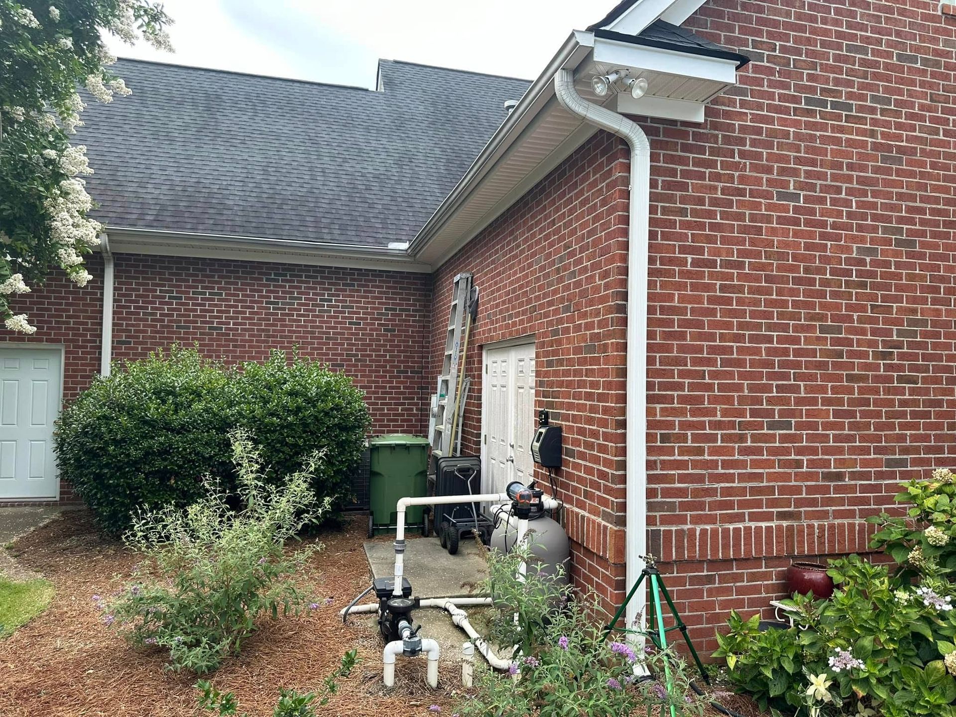 Brick house side yard with pool equipment, bushes, and a garden hose near a white door.