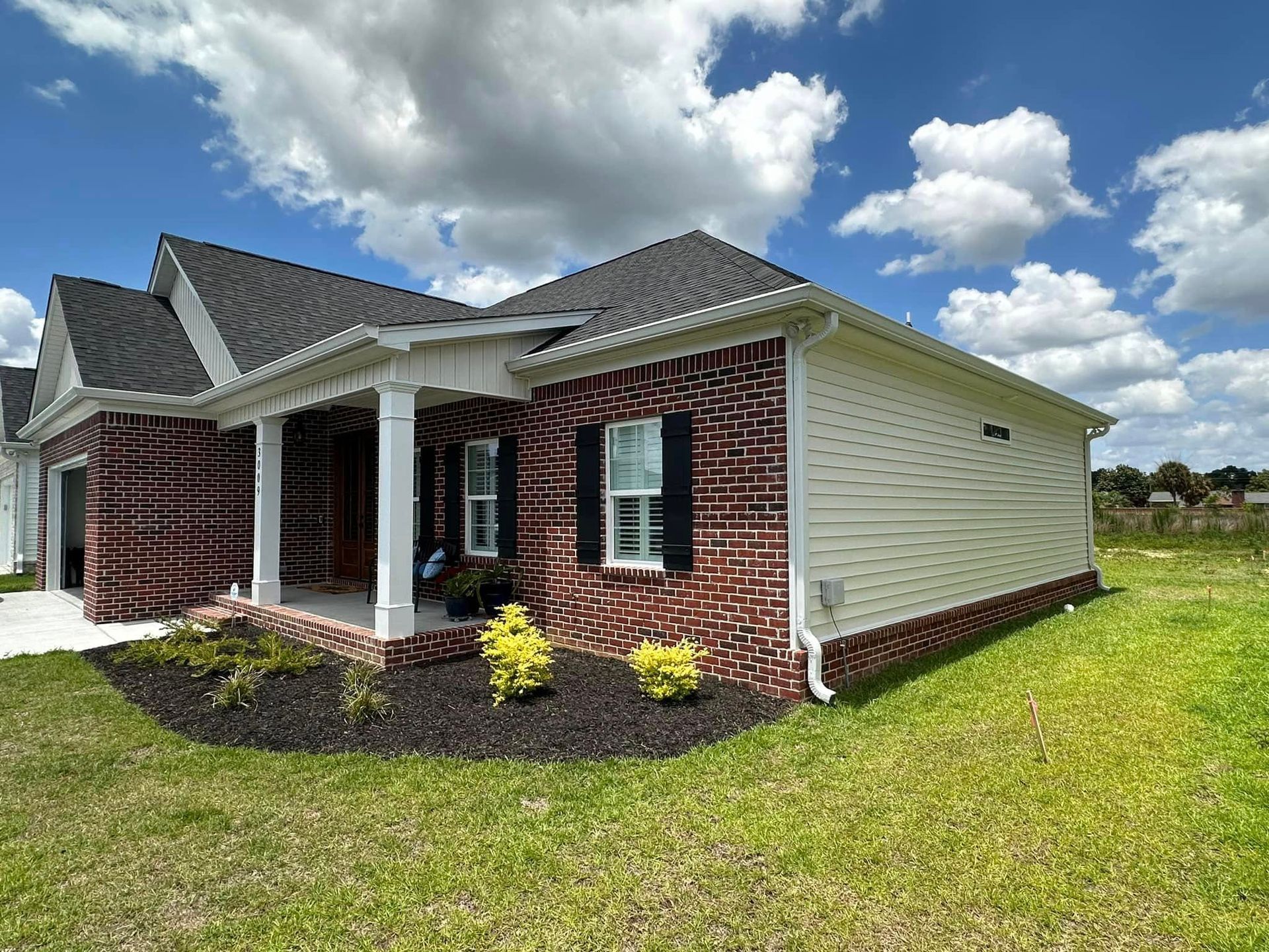 Single-story brick-and-siding house with a front porch, green lawn, and cloudy blue sky