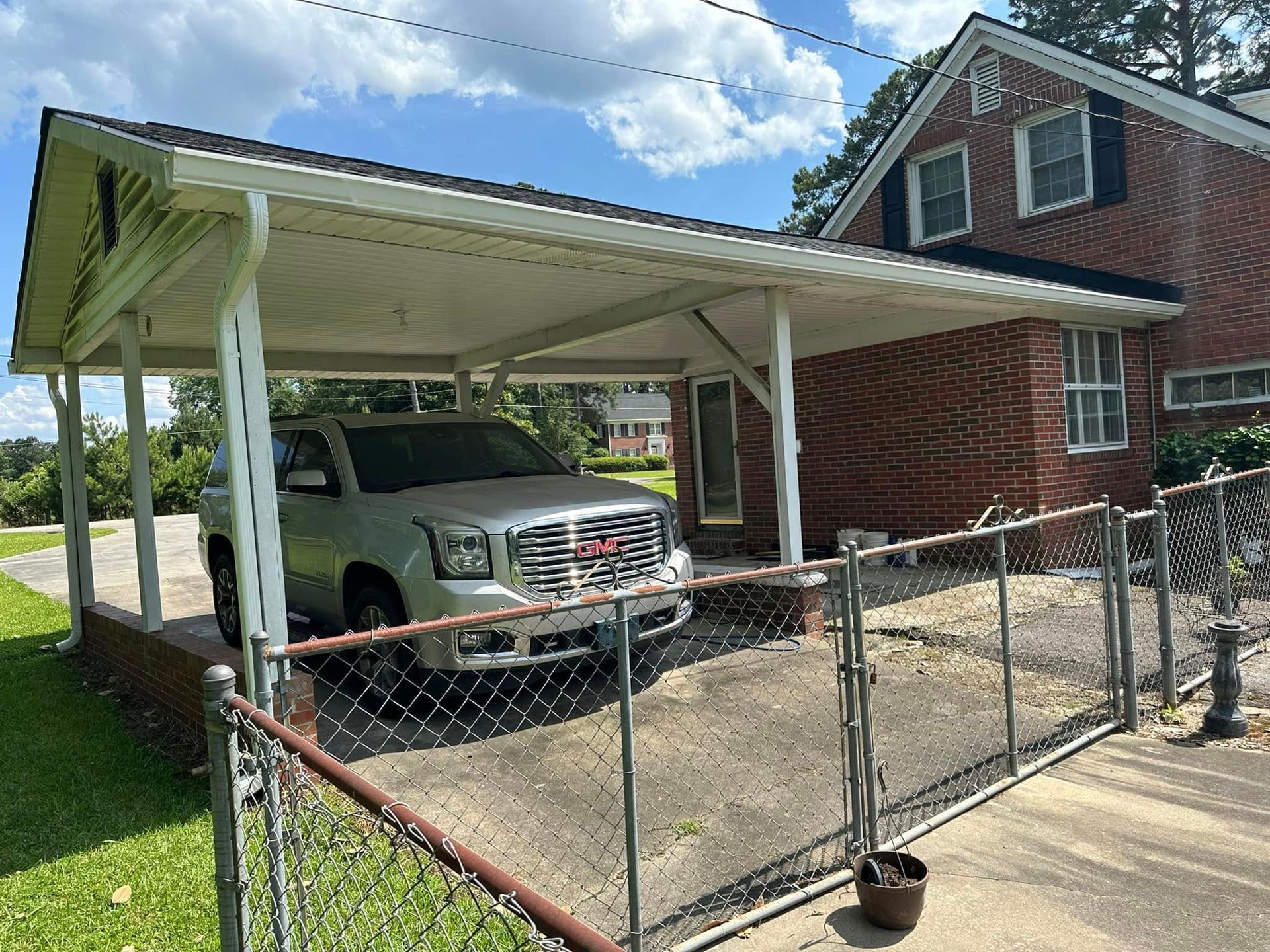 Carport beside a brick house with a white truck parked under it and a chain-link fence in front.