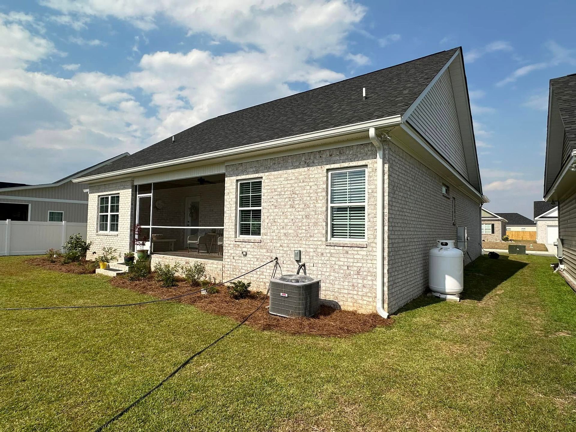 Single-story light brick house with black roof, front porch, and a grassy yard under a partly cloudy sky
