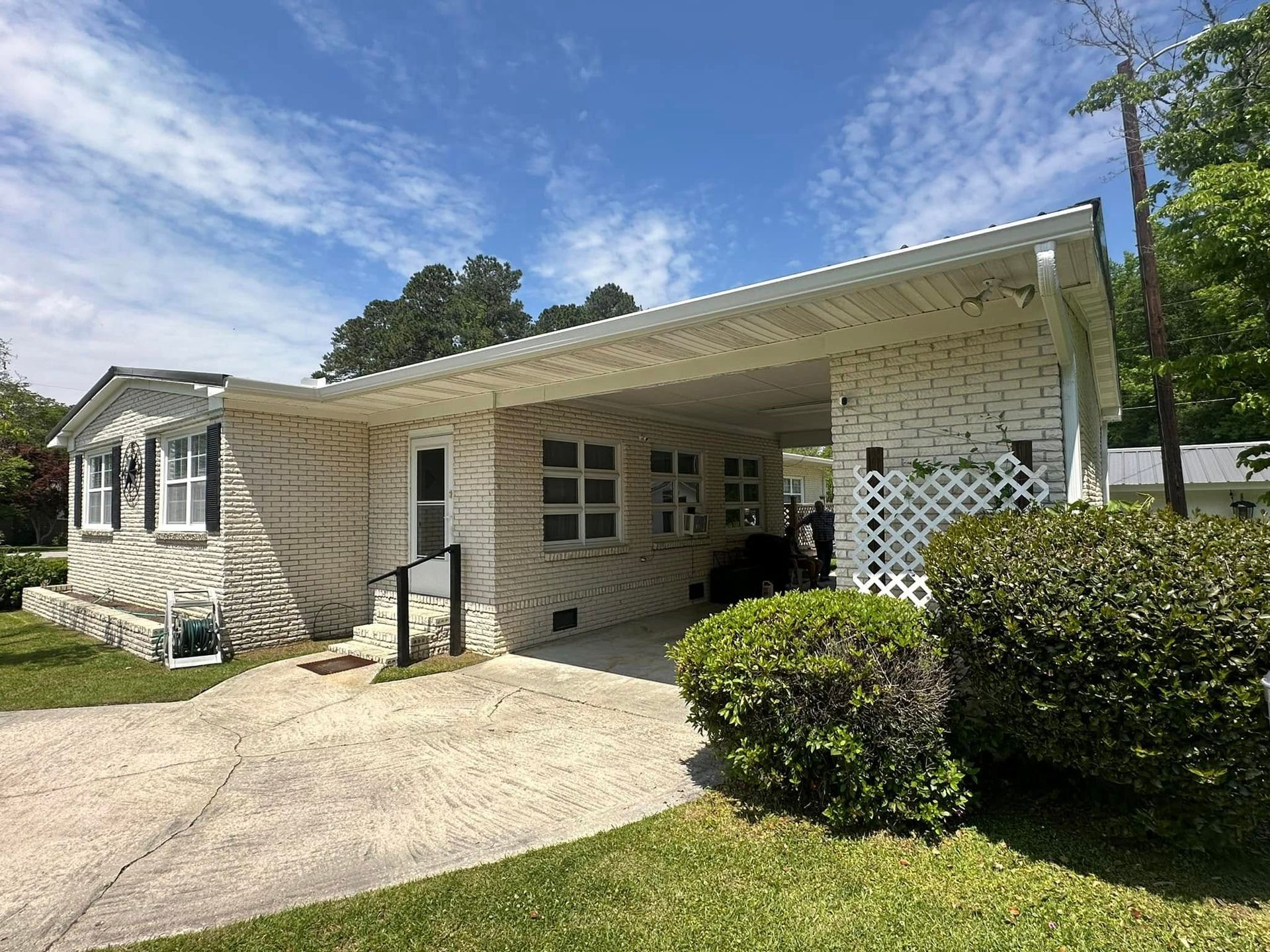White brick house with a carport, driveway, and trimmed bushes under a blue sky