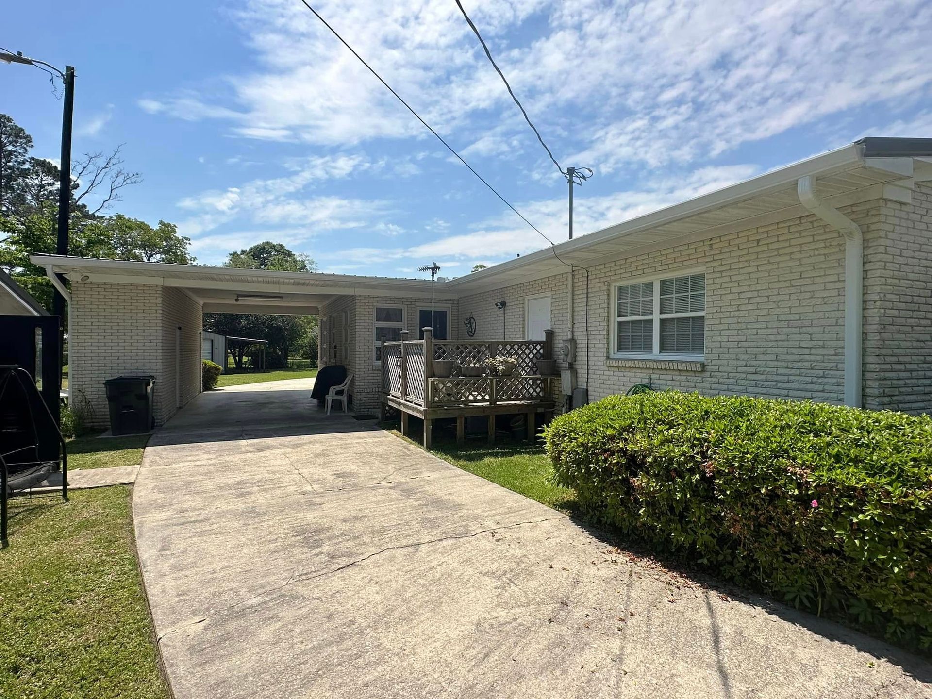 Residential driveway beside a white brick house, with a wooden deck, hedge, and carport under a blue sky