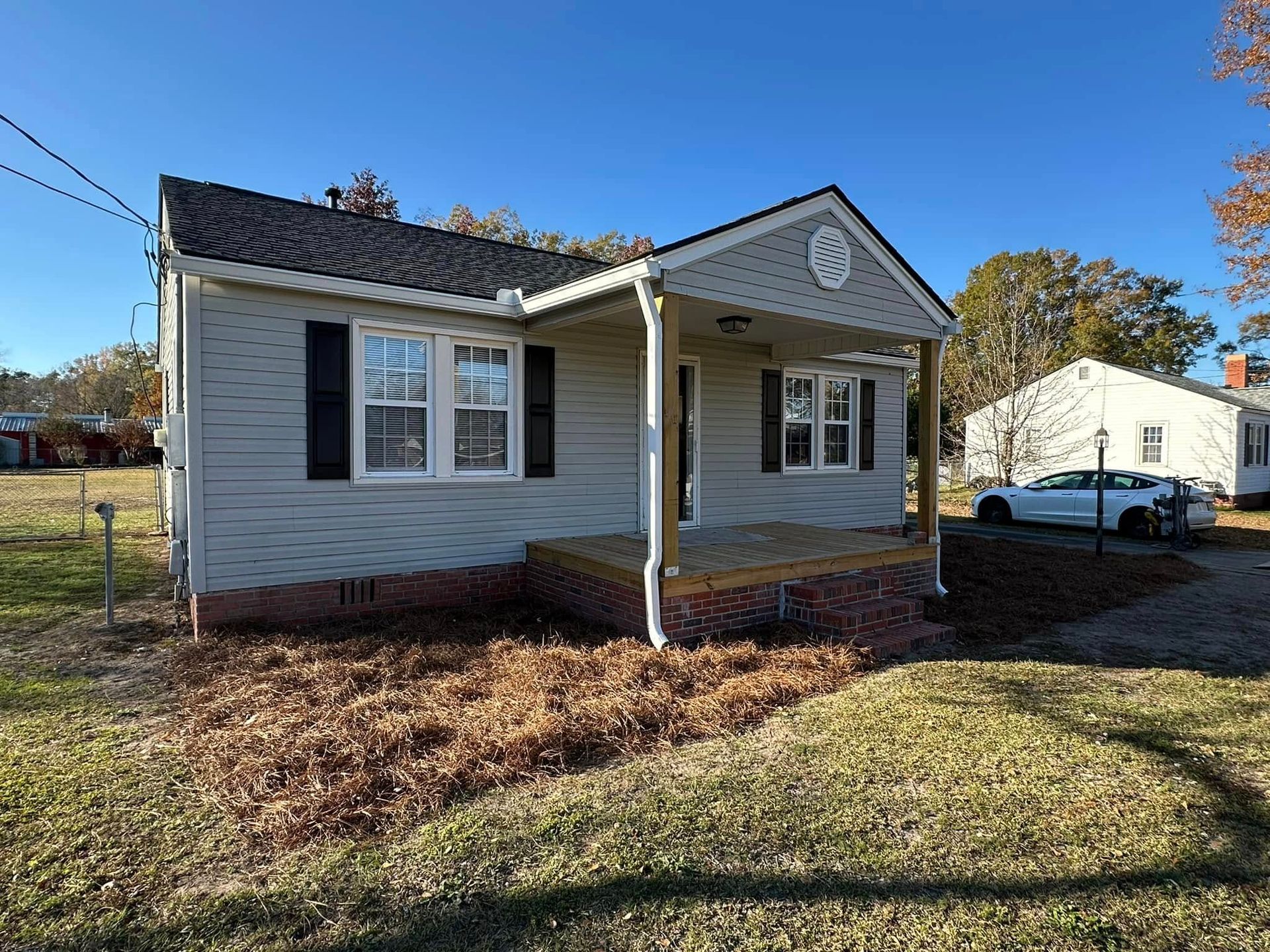 Small white house with a front porch, black shutters, and a car parked beside it on a sunny day