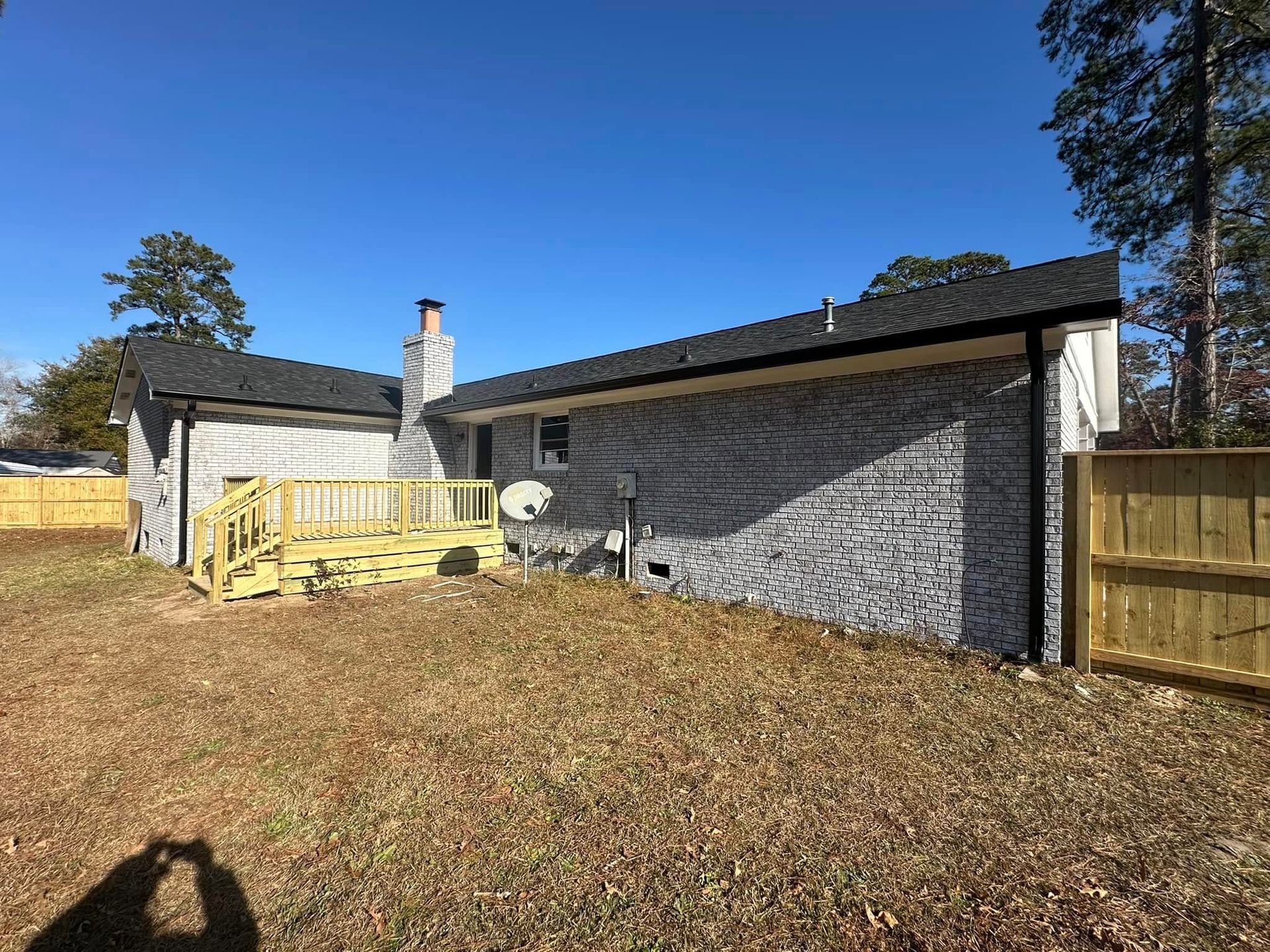 Single-story gray house with wooden deck, fenced yard, and clear blue sky