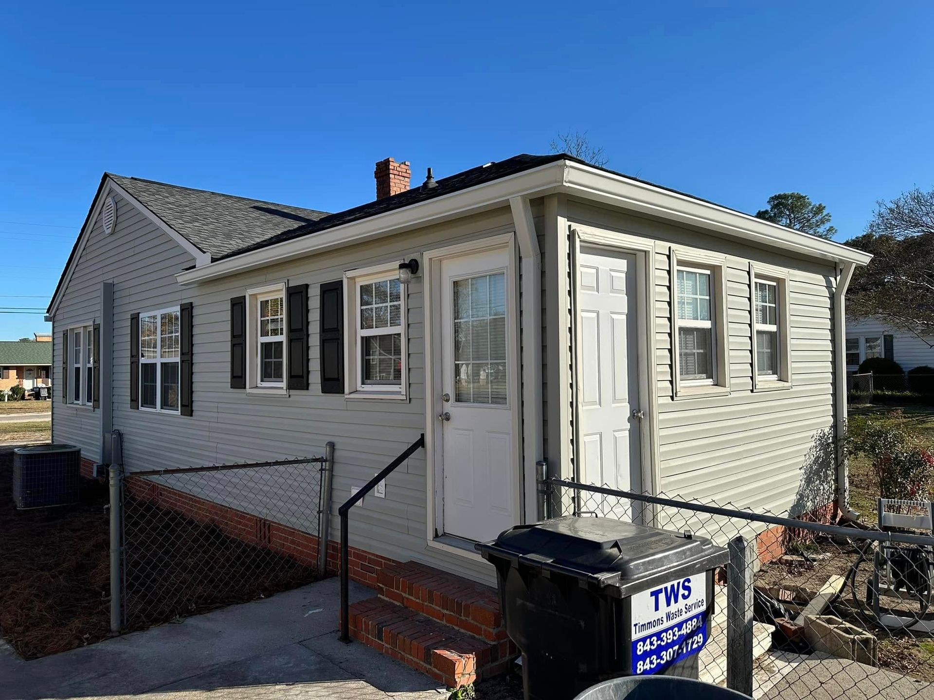 Small beige house with white trim, front steps, and a chain-link fence under a clear blue sky
